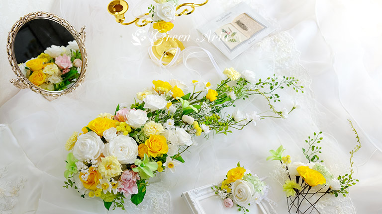 白い丸いバラと黄色のバラ薄黄色のバラグリーンのランなどの入った丈の長いウエディングブーケがドレスの上におかれている。周りにヘアパーツやブートニア、鏡が置かれている。